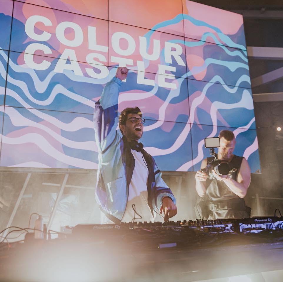 Colour Castle, also known as Christian Pillot, with a fist in the air at the decks in front of a large Colour Castle screen